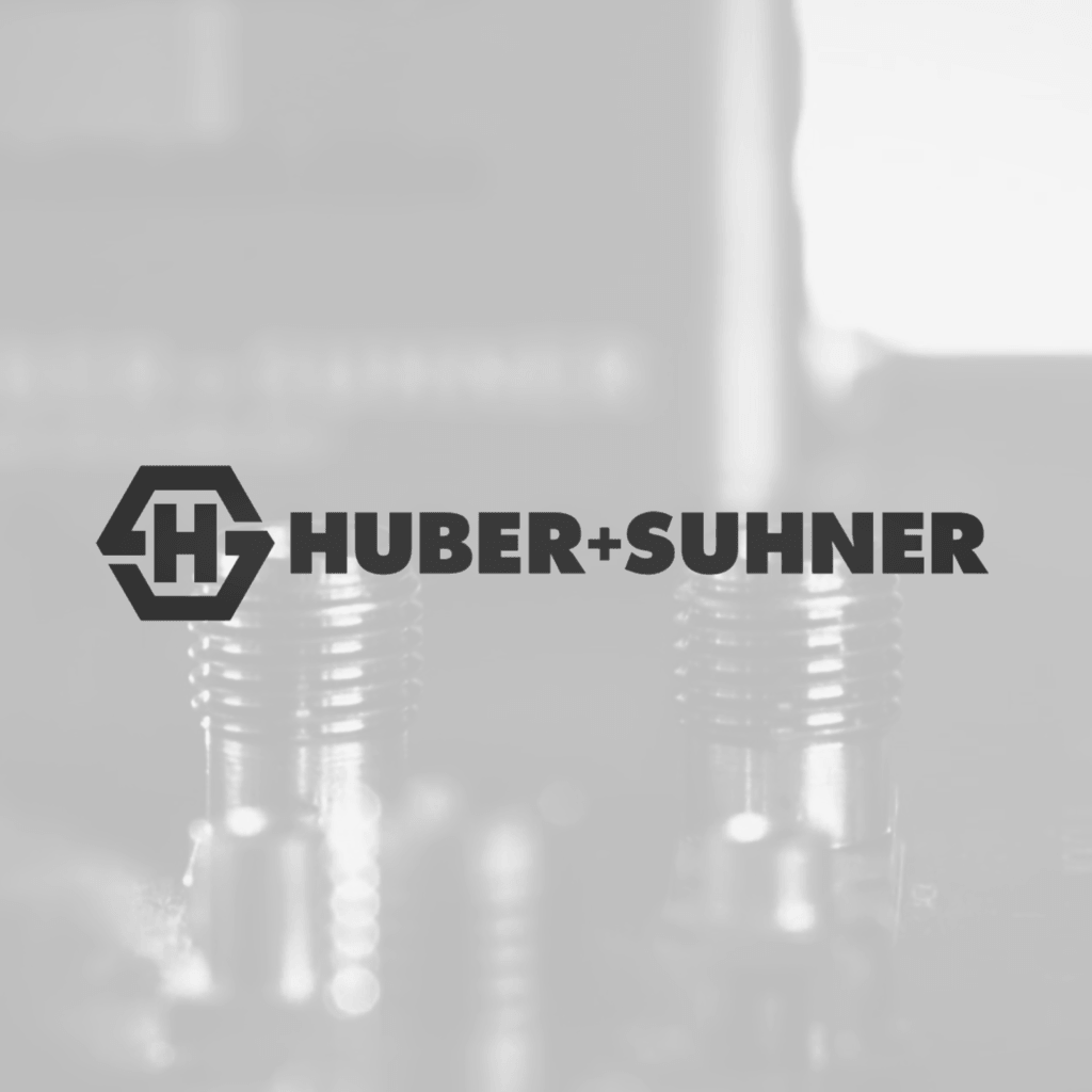 Logo mit dem Schriftzug 'HUBER+SUHNER' und einem stilisierten Hexagon mit den Buchstaben 'H' und 'S', im Vordergrund unscharfe metallische Bauteile