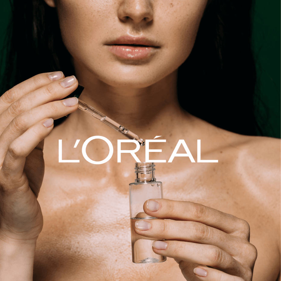 Hände halten eine Pipette und eine kleine Glasflasche vor nackter, glänzender Haut mit dem Schriftzug 'L'ORÉAL'