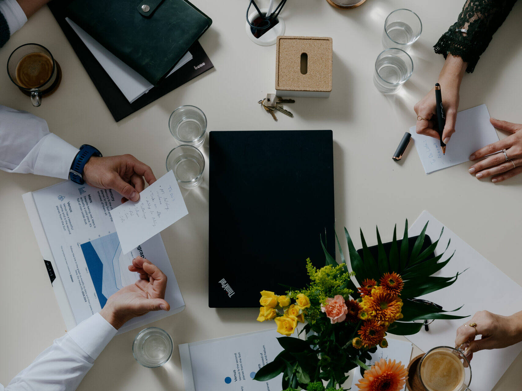 Ansicht eines Besprechungstisches mit mehreren Händen, Laptops, Dokumenten, Blumenstrauß, Gläsern, Obstschale und Lautsprecher