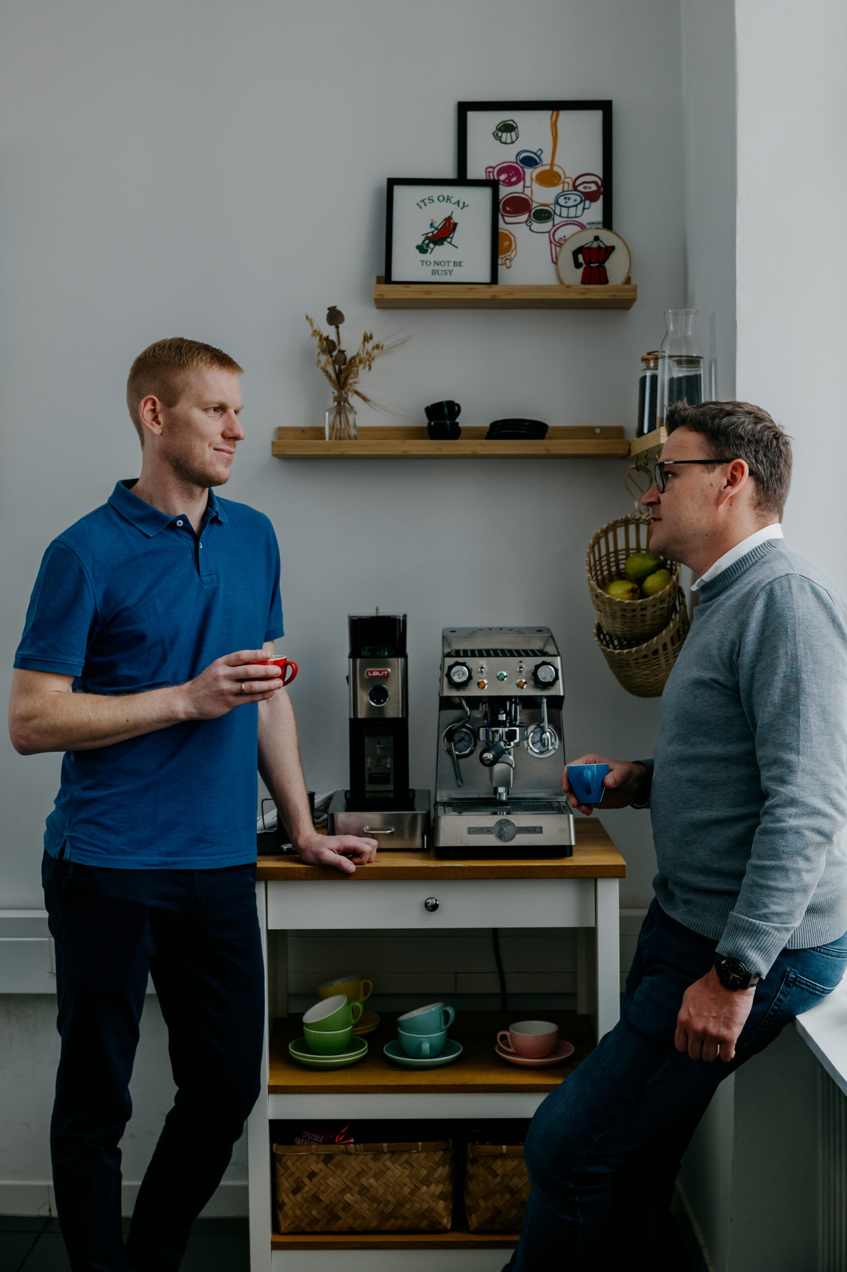 Zwei M&auml;nner stehen in einer K&uuml;che vor einer Kaffeemaschine und halten Tassen in der Hand