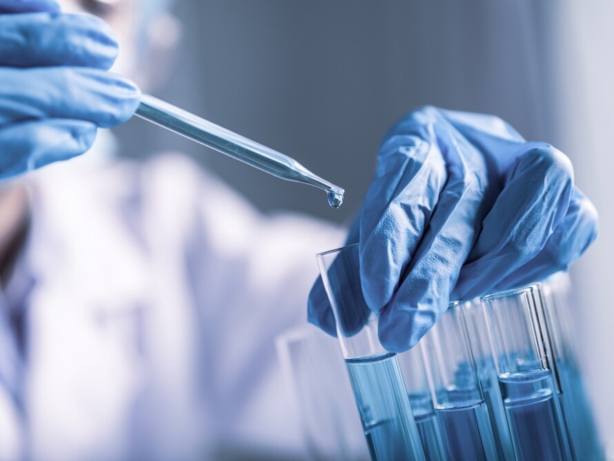 Hand mit blauer Schutzhandschuh hält Pipette über Reagenzgläsern mit blauer Flüssigkeit