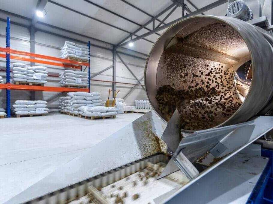 Industrielle Maschine mit rotierender Trommel und Förderband in einer Lagerhalle mit Regalen und gestapelten Säcken