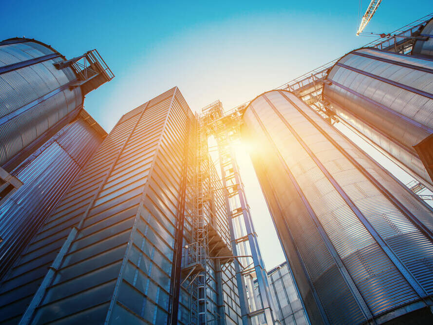 Blick von unten auf mehrere hohe silberne Industriesilos mit Sonnenlicht, das zwischen ihnen hindurchscheint